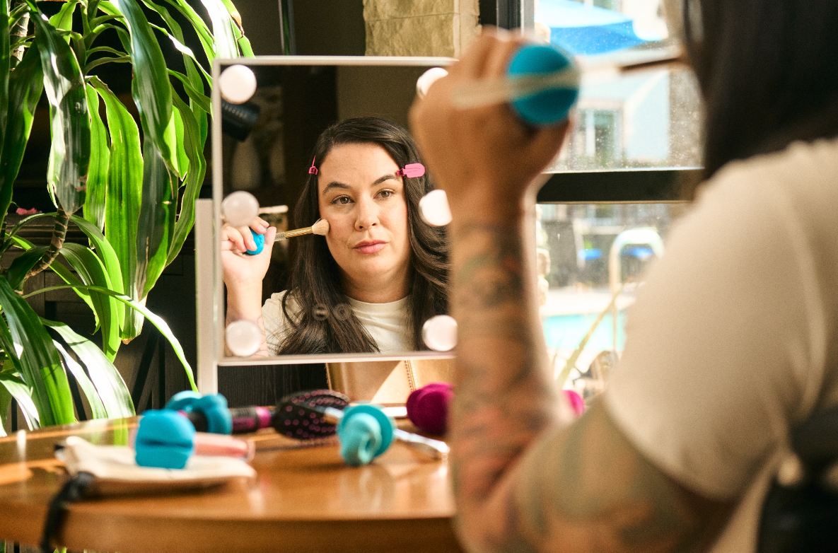 a girl with brown hair is doing her makeup with a blue medium side grip holding a makeup brush as she puts on blush. She is looking in a mirror as she uses the grips. there is a table with more beauty products and grips in front of her.